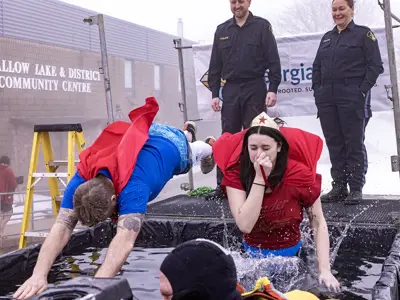 Two people dressed as superheroes in colorful costumes, one man dressed as superman with his arms stretched out like he is flying as he dives into a bin of freezing cold water, one woman dressed as Wonder Woman with a crown on doing a cannonball into the bin plugging her nose. Both are wearing capes. Two police officers standing on scaffolding watching them jump in while smiling. Two dive team members from the fire department inside the bin filled with water to help them out. 